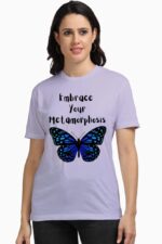 A person wearing the Stylish and Empowering Women T-Shirt from Looga, featuring a blue butterfly and the message "Embrace Your Metamorphosis" on a light purple background, exudes inner transformation against a plain white backdrop, ready for their personal looga moment.