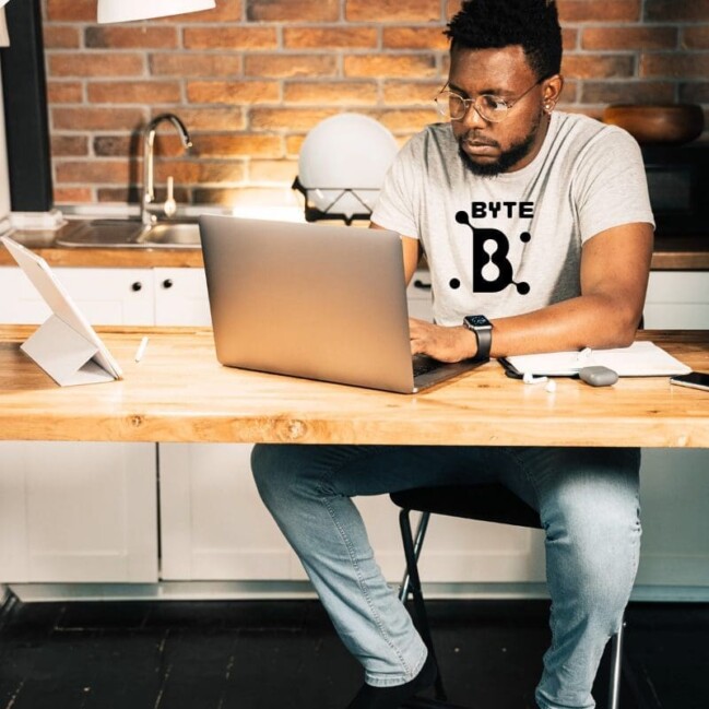 A man sits at a wooden table in a cozy kitchen, working on his laptop. Sporting glasses and a gray t-shirt with BYTE printed on it, he dives into his work. A tablet, notebook, and pen accompany him as the soothing ambiance of modern lighting against the brick wall creates a calming looga.