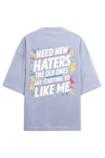 The image showcases a light blue Looga Premium Oversized T-Shirt, 100% cotton and bio-washed, featuring the statement Need new haters, the old ones are starting to like me, surrounded by colorful lightning bolts and stars for emphasis.