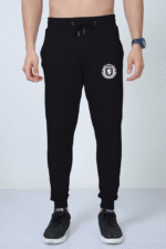 A person epitomizing style and comfort stands against a white background, showcasing the Premium Joggers for Men - Comfort Meets Style at Looga. These black joggers feature a crest on the left thigh and are paired with black sneakers and a blue wristwatch. They include an adjustable drawstring waistband to ensure the perfect fit.