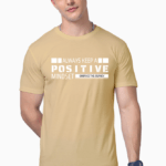 A person in a beige Unisex Supima T-Shirt with Always Keep a Positive Mindset, Embrace the Journey printed on it stands with short dark hair, embodying the laid-back Looga style and posing with one hand in their pocket.