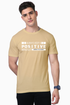 A person in a beige Unisex Supima T-Shirt with Always Keep a Positive Mindset, Embrace the Journey printed on it stands with short dark hair, embodying the laid-back Looga style and posing with one hand in their pocket.