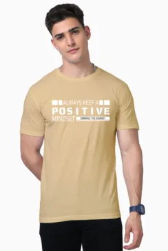 A person in a beige Unisex Supima T-Shirt with Always Keep a Positive Mindset, Embrace the Journey printed on it stands with short dark hair, embodying the laid-back Looga style and posing with one hand in their pocket.