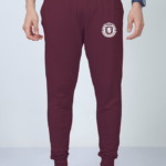 A man wearing Looga's Premium Joggers for Men - Comfort Meets Style, in a sophisticated burgundy color with a white emblem on the upper thigh and an adjustable drawstring, stands against a gray background. Completing his look are black sneakers with white soles and a sleek blue watch.