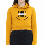 A person wearing a yellow cropped hoodies with SMILE in large letters, a crossed-out word above it, and a smiley face graphic below. They have long hair and are standing against a plain background.