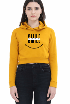 A person wearing a yellow cropped hoodies with SMILE in large letters, a crossed-out word above it, and a smiley face graphic below. They have long hair and are standing against a plain background.