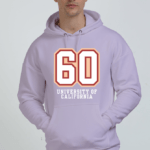 Wearing a stylish Lavender hoodie with 60 and University of California on the front, a person with short dark hair confidently stands against a plain background, effortlessly showcasing the perfect mix of heavyweight hoodies and style.