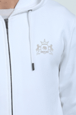A person is wearing a white, thick embroidered hoodie featuring a barely visible subtle emblem on the chest with a crown, two lions, and the text SINCE 2023 lightly embossed against the fabric.