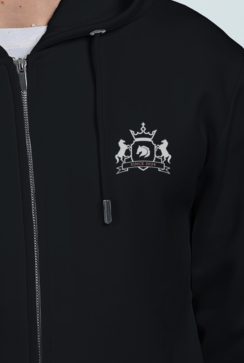 A person wears a thick, embroidered black hoodie with a silver emblem on the chest featuring two horses, a shield, and a crown beneath SINCE 2013. The hoodie includes a zipper and drawstrings.